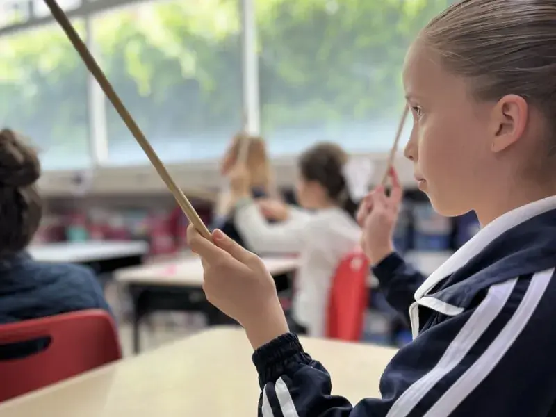 Estudiante sostiene vara mientras participa en actividad artística en aula de colegio con compañeros de clase